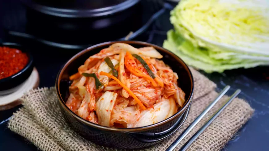 Mangkuk berisi kimchi segar dari kol yang dipotong dan dibumbui cabai merah, diletakkan di atas alas goni dengan latar dapur tradisional.