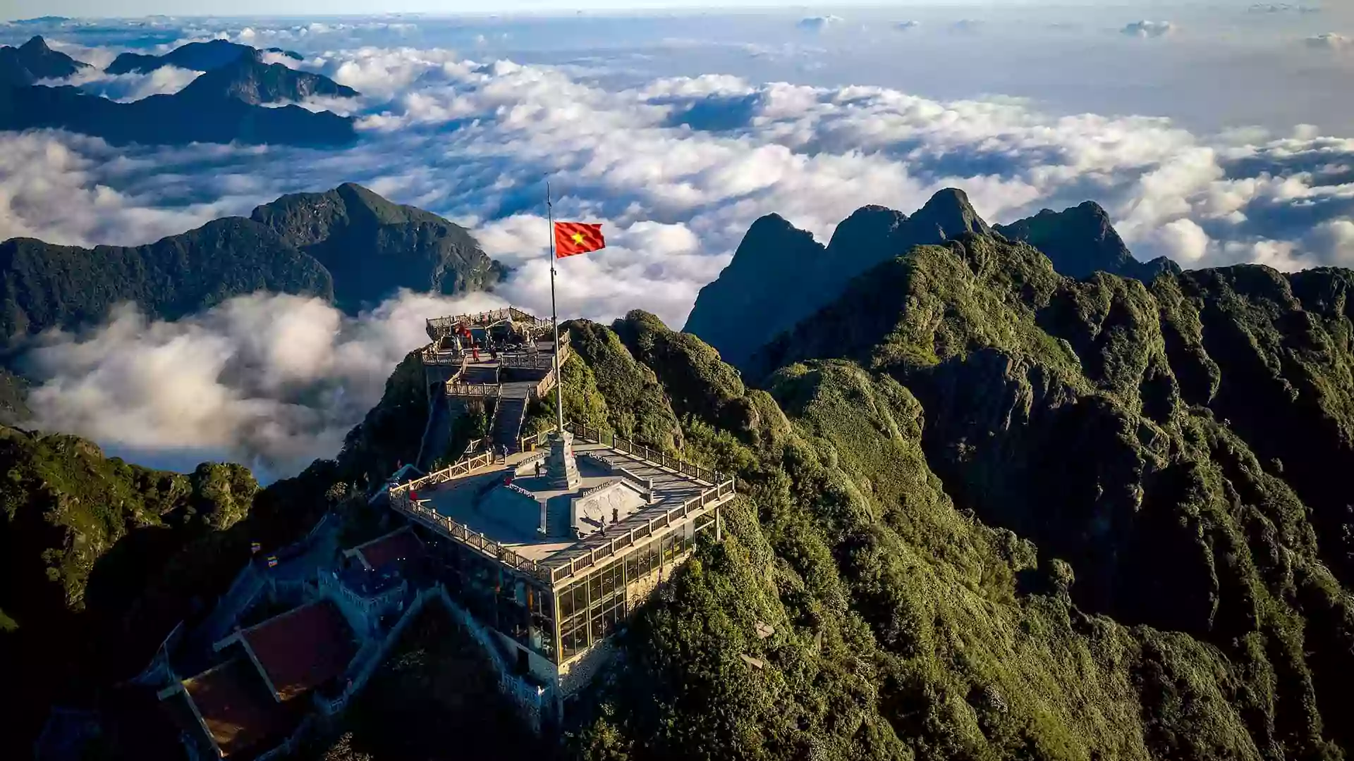 Pemandangan pegunungan Sapa dengan lautan awan, puncak berbatu hijau, dan bendera Vietnam berkibar di area pandang yang tinggi.