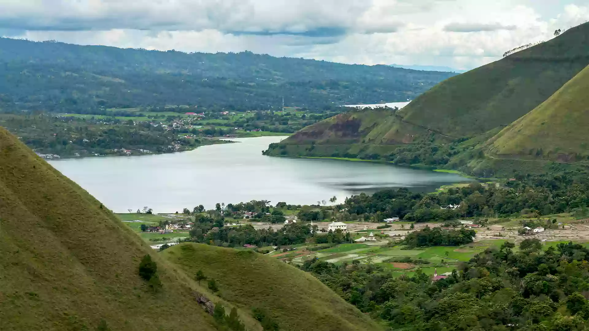View Danau Toba dari Bukit Holbung dengan perbukitan hijau, danau luas, dan desa kecil di sekitarnya.