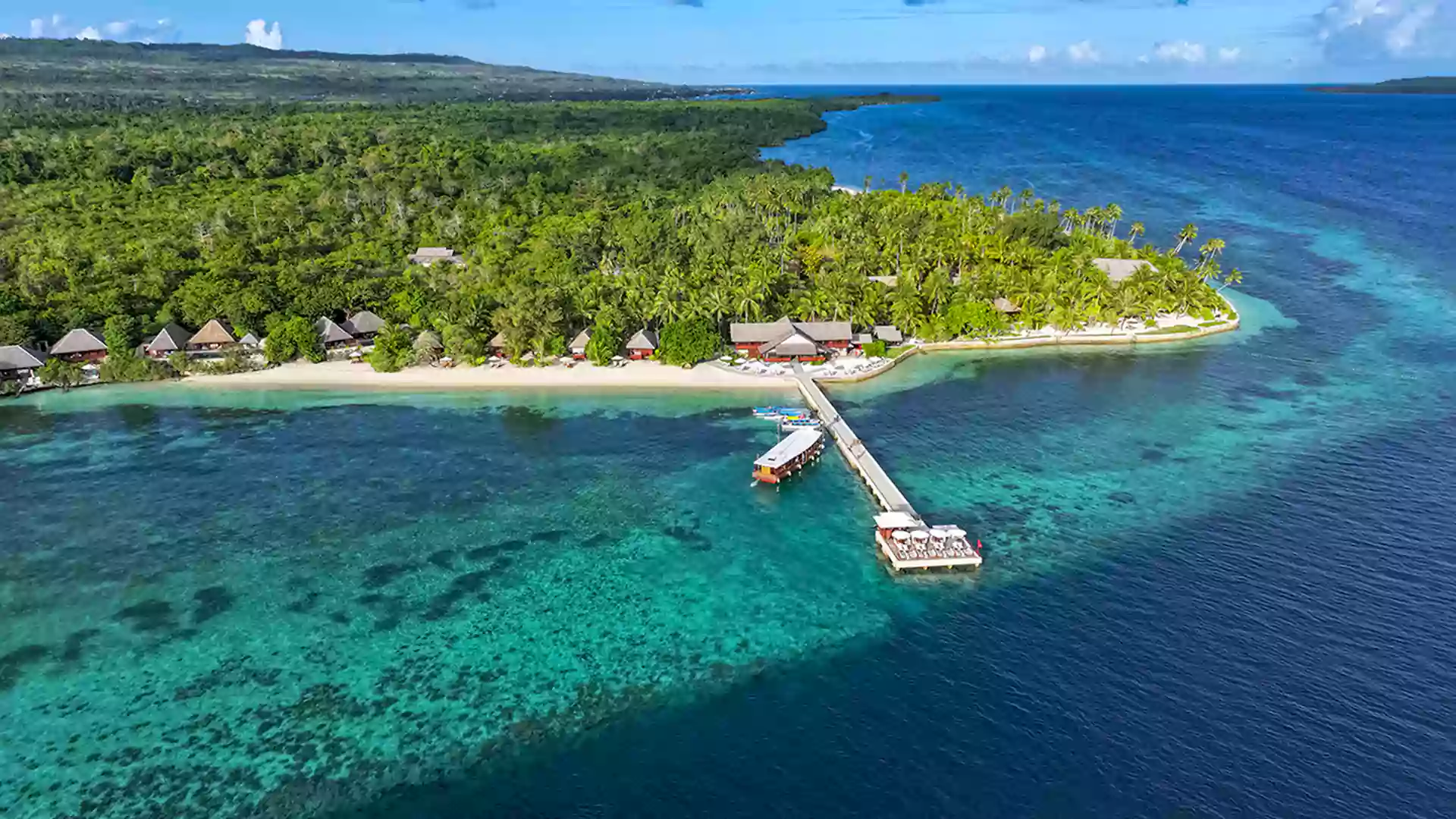 Pemandangan udara pulau tropis Wakatobi dengan air laut jernih biru kehijauan, dermaga kayu, dan resort di tepi pantai.